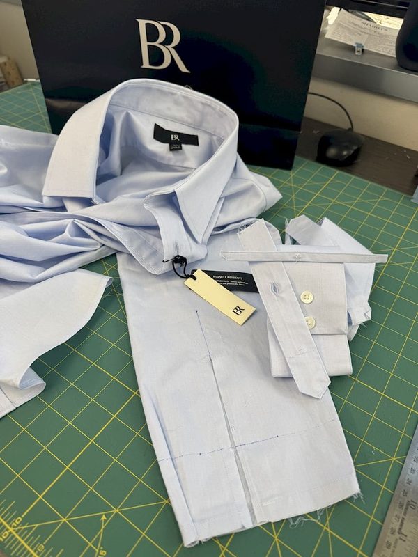 A pale blue shirt on the cutting mat with the collar removed, seam lines marked in chalk and the cuff pinned.